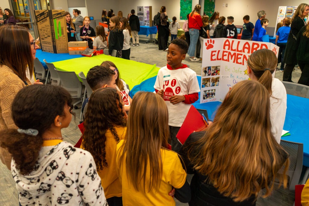 A student speaks with other students and a teacher in front of a display poster.