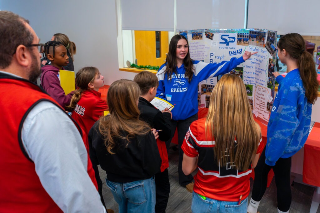 A student points to a display poster while speaking with students and teachers.