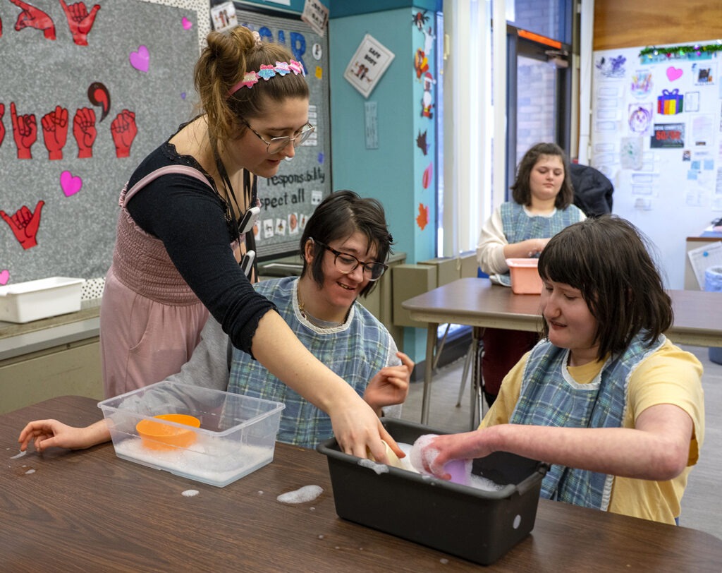 two students interact with teacher during water play table